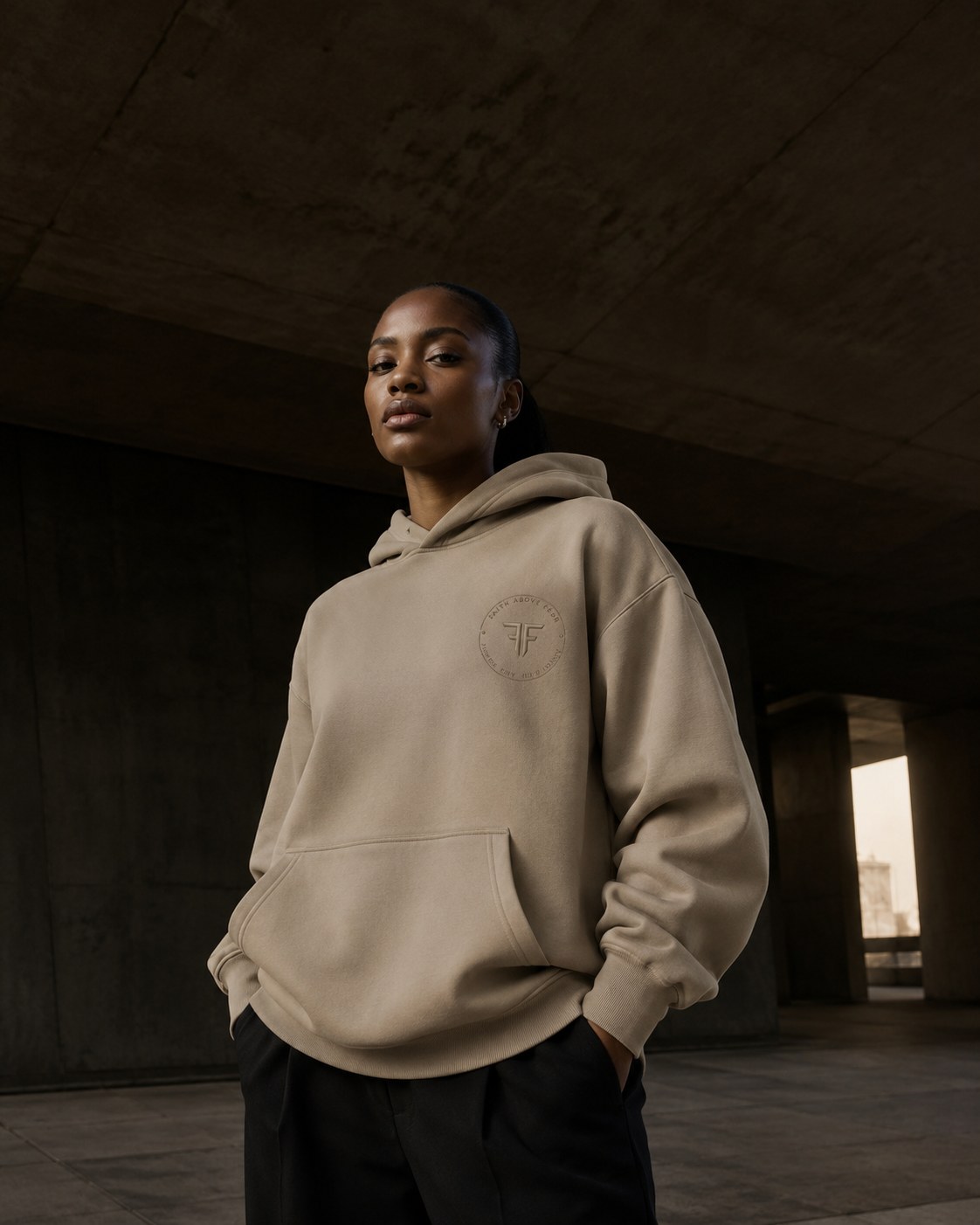 Woman wearing the sand Faith Above Fear seal hoodie with black trousers, standing under a concrete underpass