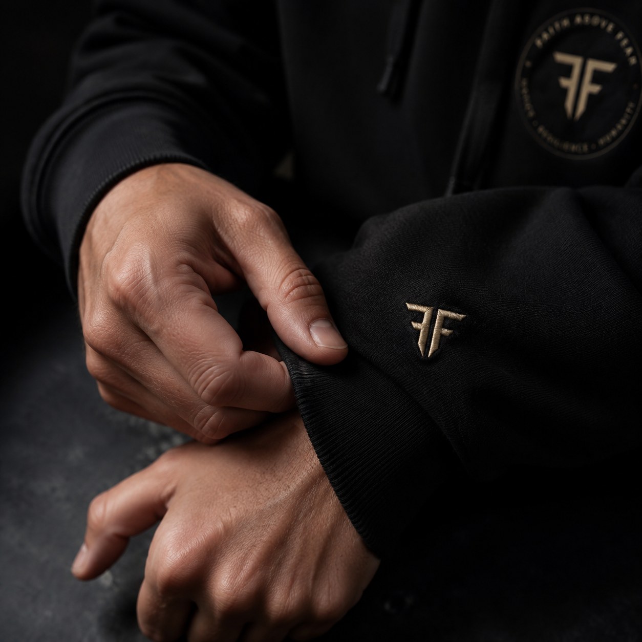 Close-up of hands adjusting a black Faith Above Fear cuff with the gold FF monogram embroidered into the fabric